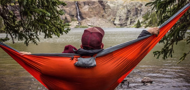 Master Your Camping Hammock Setup for Ultimate Outdoor Comfort Person relaxing in a red and gray hammock by a mountain lake, surrounded by pine trees, with a waterfall and rocky cliffs in the background – peaceful outdoor camping scene.