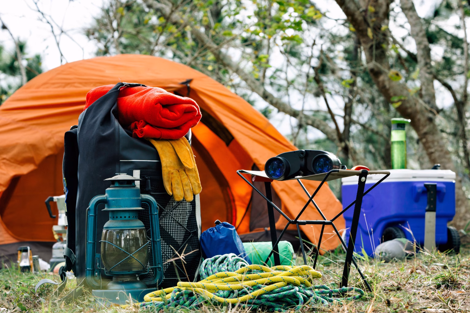 Your Ultimate Guide to Essential Camping Equipment: Gear Up for Adventure Essential camping gear setup with orange tent, backpack, lantern, gloves, rope, binoculars, cooler, and supplies in a forest campsite – outdoor adventure preparation