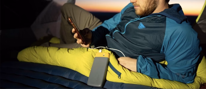 The best Portable Power Banks for Camping 2025 Young man relaxing inside a camping tent at sunset, using his smartphone while charging it with a power bank placed on a sleeping bag, wearing a blue jacket and mesh cap – perfect outdoor camping scene