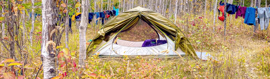 Backcountry camping tent set up in a colorful autumn forest with clothes hanging to dry on a line between trees – peaceful fall outdoor adventure scene