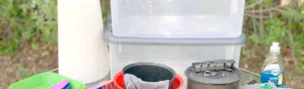 Camping Cooking Utensils List: Your Essential Guide to Outdoor Culinary Adventures Essential camping kitchen gear on an outdoor table including cast iron skillets, mugs, plates, bowls, utensils, scissors, can opener, cutting boards, dish soap, paper towels, aluminum foil, and plastic storage bins in a forest setting.