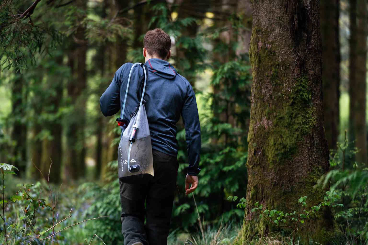 Man hiking through a lush green forest carrying an MSR Autoflow XL gravity water filter bag over his shoulder for outdoor water purification.